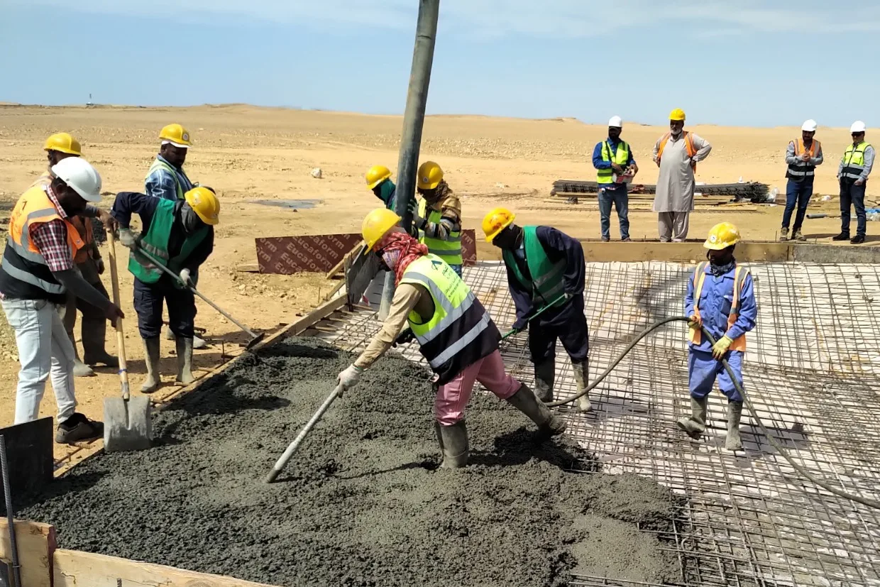 Crew placing and vibrating concrete over reinforcement mesh on a desert construction site while engineers look on